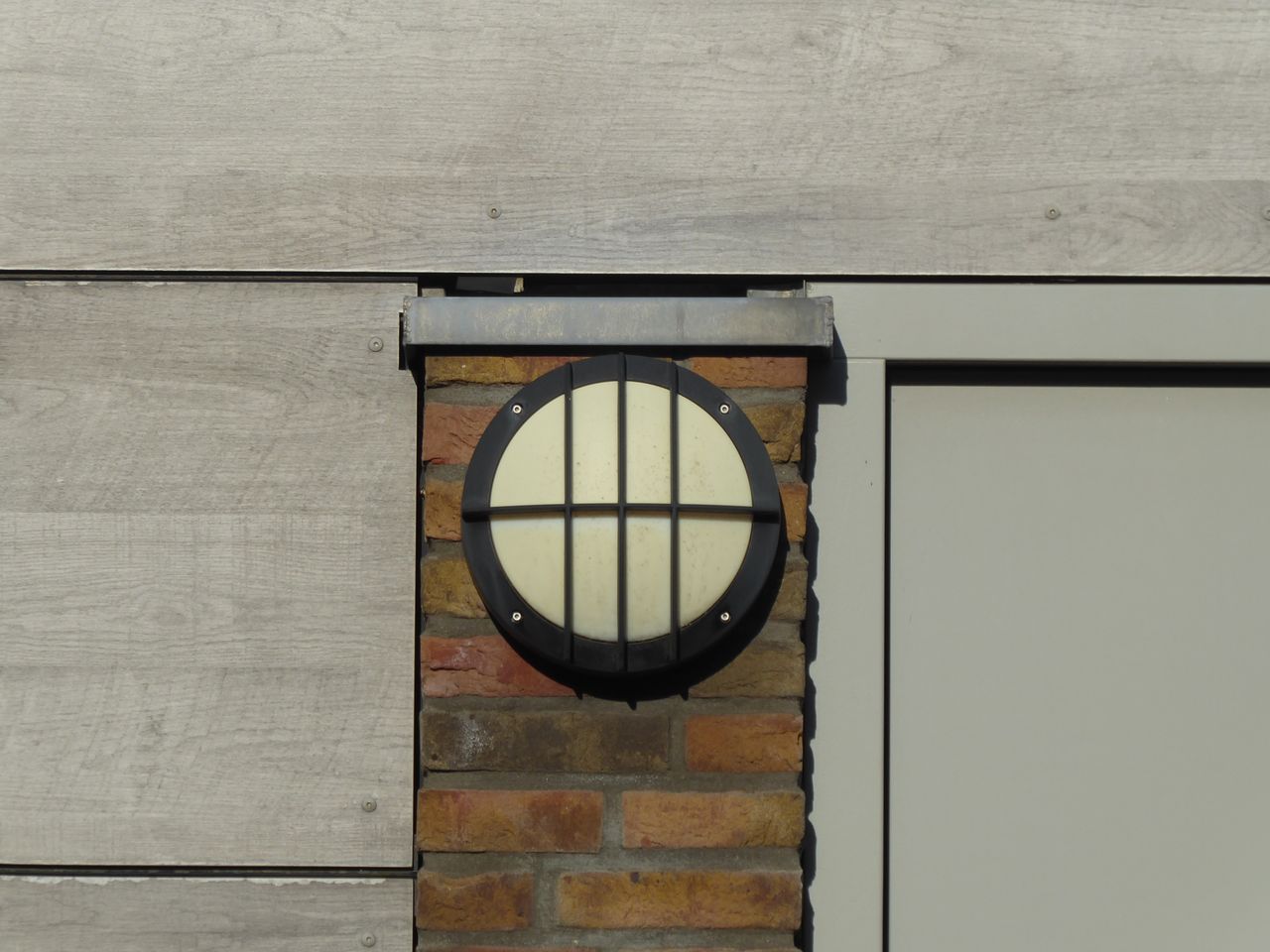 A round industrial lamp mounted on a red brick wall with wooden siding.
